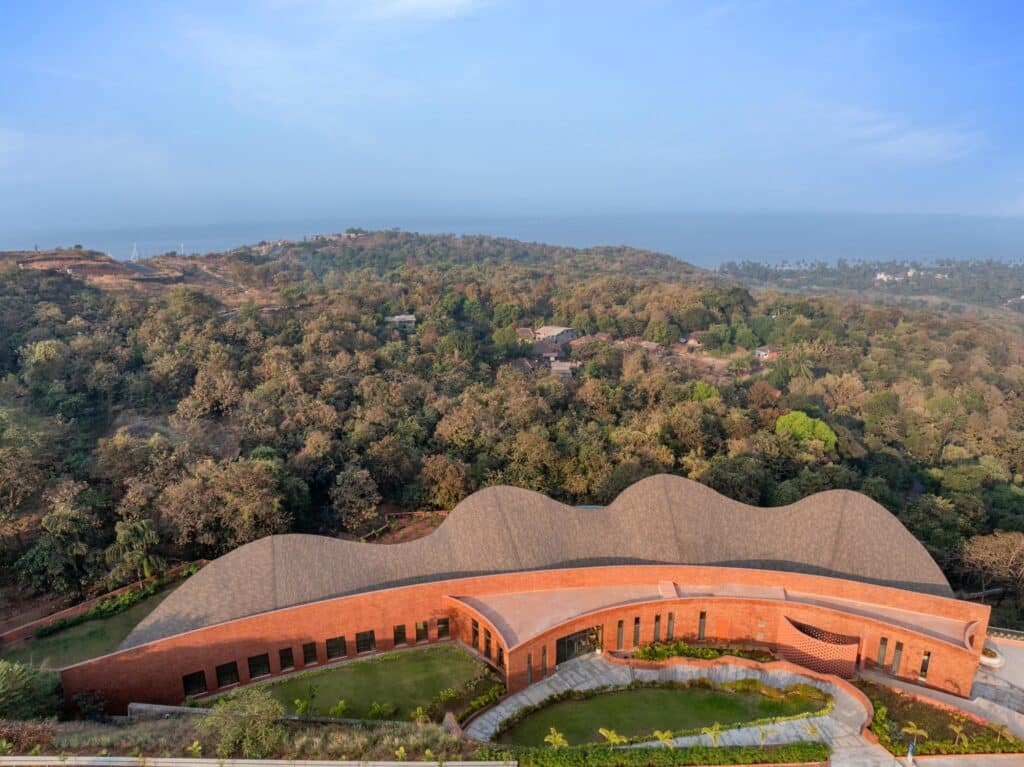 Aerial view of CREST NINE Community Center featuring a curved red laterite stone facade and undulating roofline against a coastal forest backdrop.