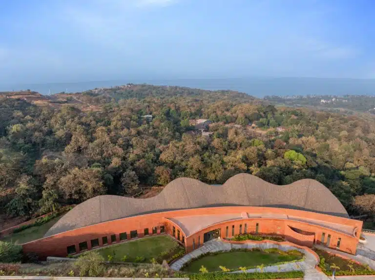 Aerial view of CREST NINE Community Center featuring a curved red laterite stone facade and undulating roofline against a coastal forest backdrop.