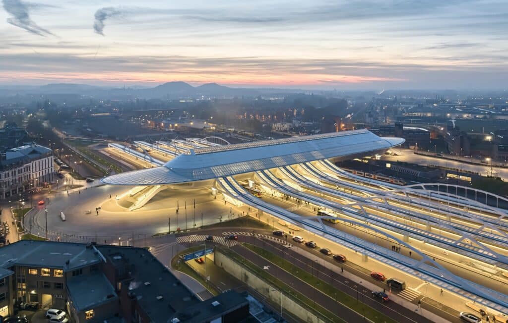 An aerial sunset view of the Mons Train Station in Belgium, showing the white steel bridge structure spanning over multiple railway tracks.