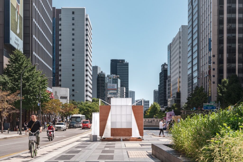 A white and wood architectural pavilion named TEUM situated on a busy city sidewalk with skyscrapers in the background.
