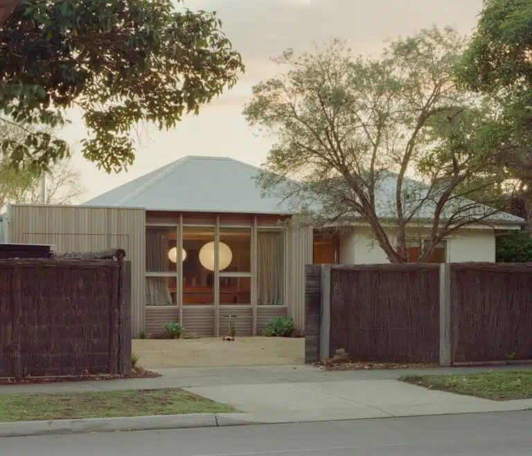 Front facade of Wood House in Barwon Heads showing timber cladding, large glass windows with warm interior lighting, and a natural brush fence.