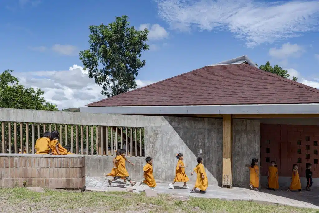 Children walking along a concrete and brick wall under a large hip roof at Sonomoro School, showing the transition between public and private spaces.
