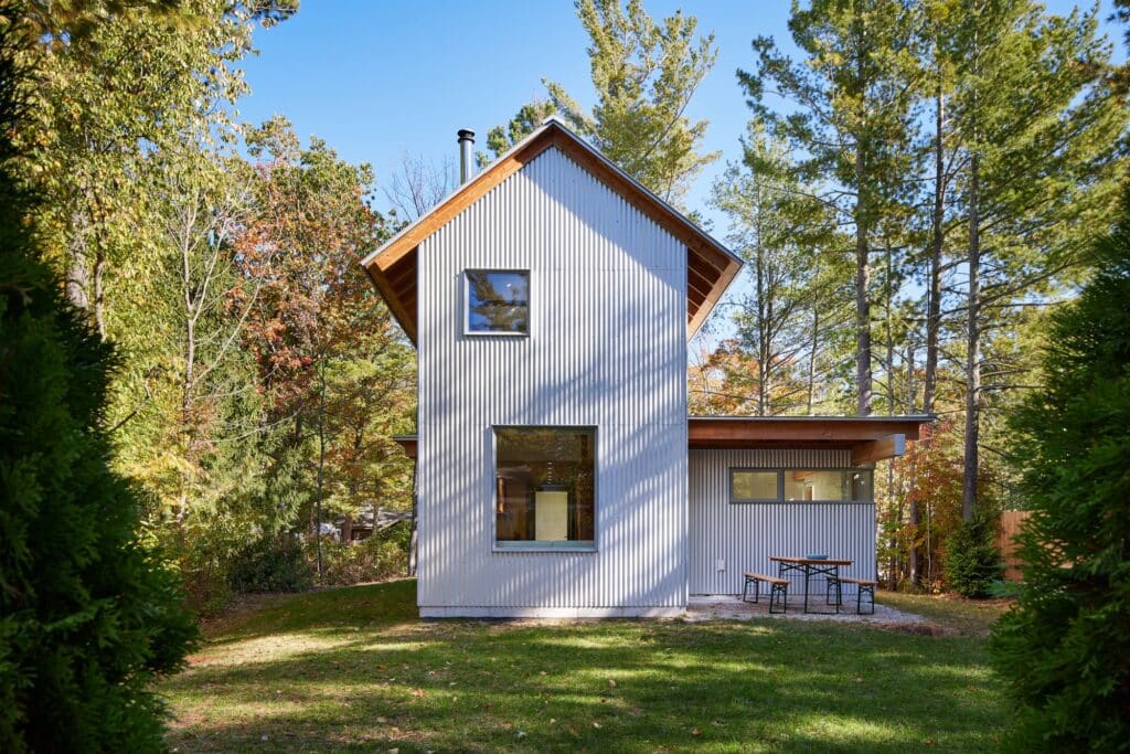 Rear facade of the Liten Hytte House with large windows and a small outdoor sitting area in a grassy clearing.