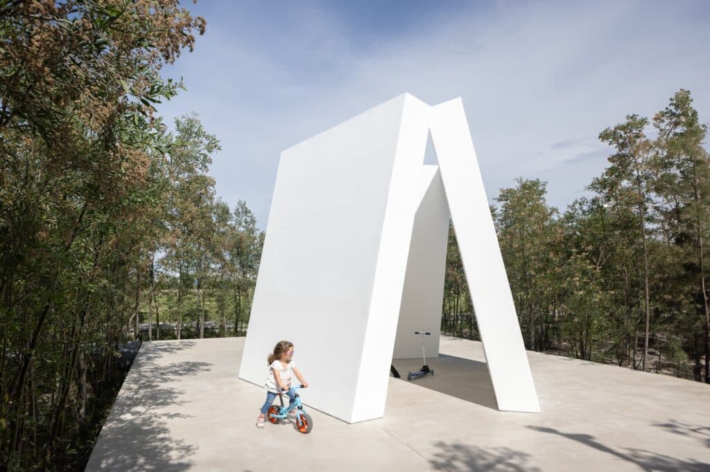 A child on a bike in front of a modern white triangular pavilion at Malba Puertos cultural center.