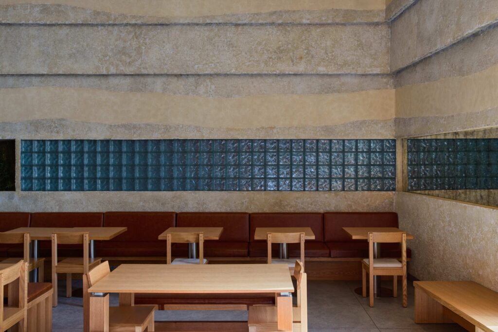 Textured interior wall with horizontal sand-like layers and a strip of blue glass bricks above wooden dining tables.