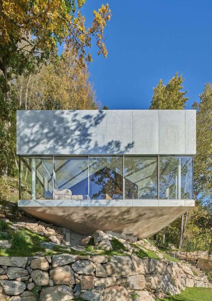 Close-up of the main cabin at Bor&aring;s Getaway House showing the galvanized steel upper half and glass lower half supported by a concrete base on rocks.