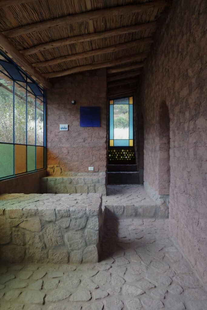 Interior corridor featuring textured adobe walls, stone seating, and a stained glass window with yellow and blue accents.