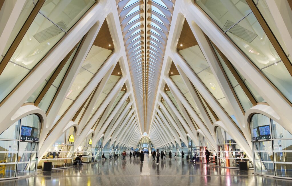 Interior perspective of the main passenger gallery at Mons Station featuring a rhythmic sequence of white triangular steel trusses and glass roofing.