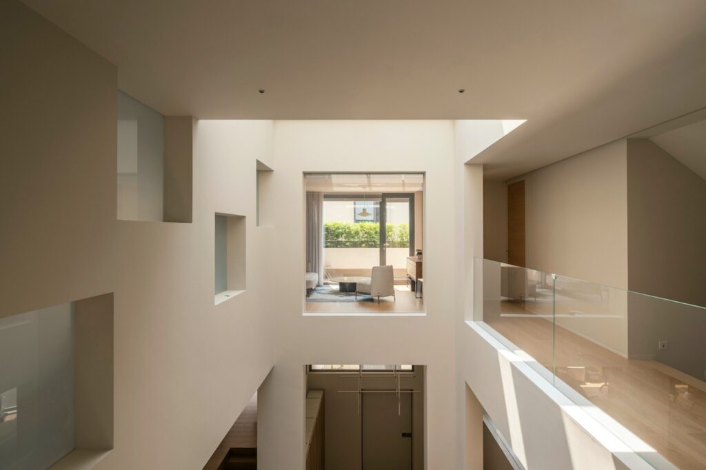 Interior perspective from an upper floor looking through internal windows towards a living area with a view of a balcony.