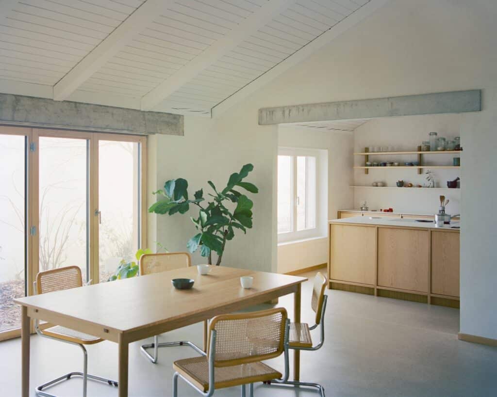 Interior view showing the visual connection between the dining table and the renovated kitchen area. Exposed concrete beams and white wood ceilings define the architecture.
