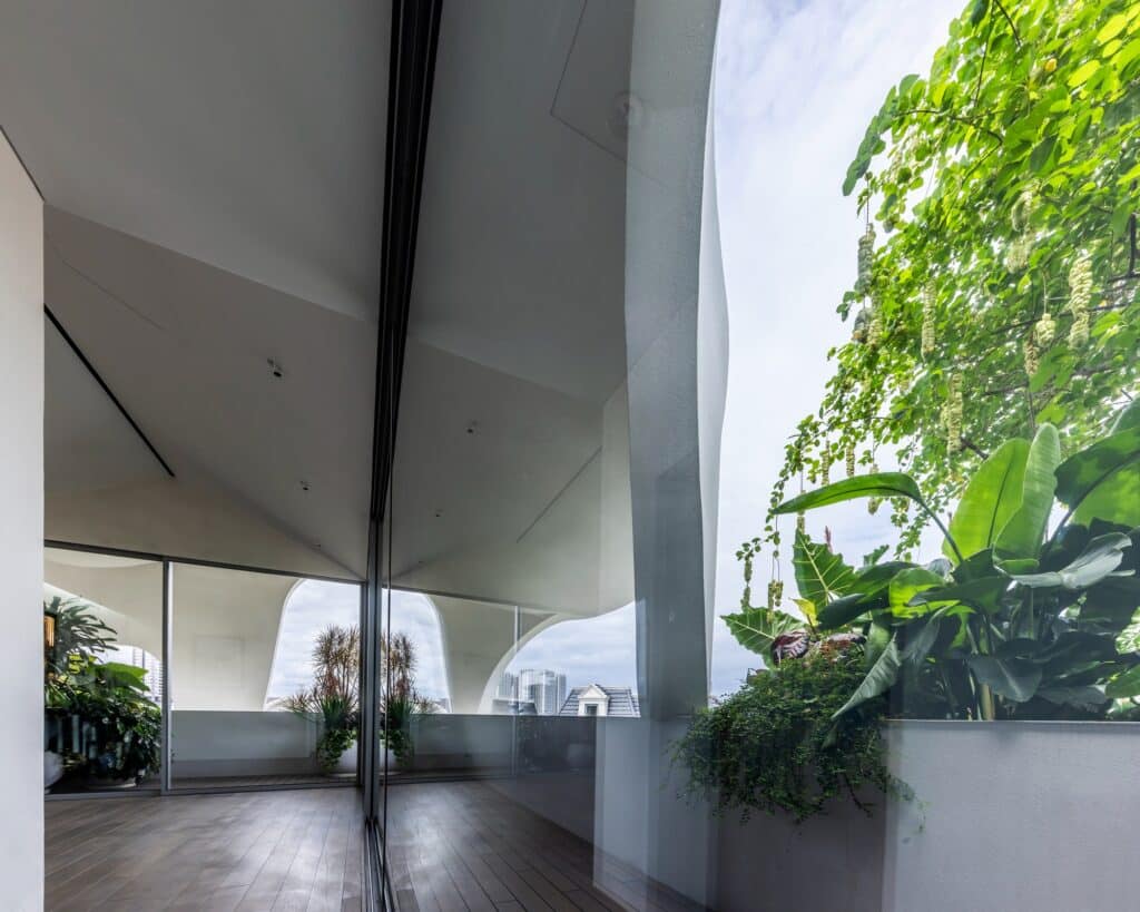 Interior view through a large glass sliding door reflecting the outdoor garden and curved white ceiling.