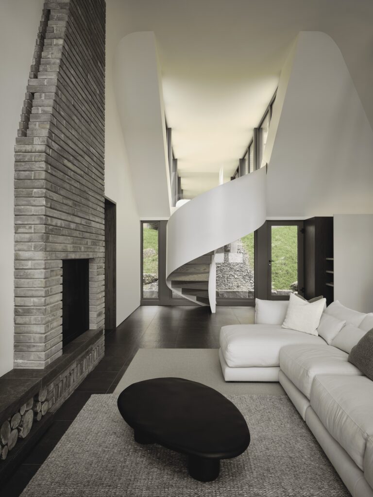 Interior of Maison de la Pointe living area featuring a modern, white sculptural spiral staircase and a large, traditional grey brick fireplace against a double-height wall.