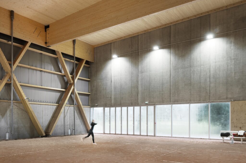 Interior view of a Sporting Vichy tennis court featuring large glulam timber beams and a raw concrete wall with industrial lighting.