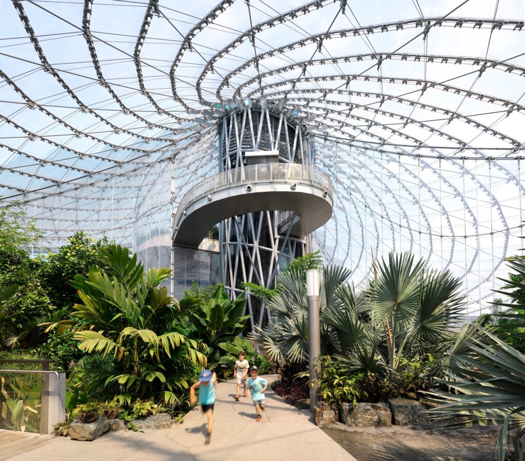 Children running through the tropical biome at The Leaf with a view of the spiral walkway and the vast cable-net roof structure.