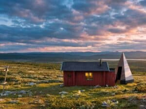 Black pyramid-shaped Klumpen cabin next to a traditional red wooden house in a vast polar tundra landscape under a cloudy sunset.