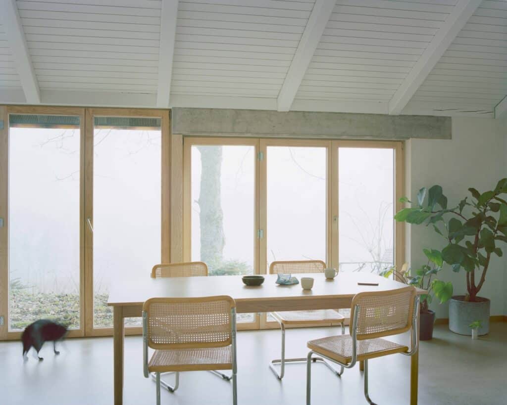 A wooden dining table and chairs set against large floor-to-ceiling wooden windows overlooking a foggy forest landscape.