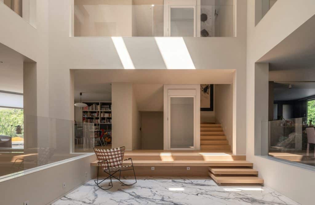 Interior view of the central light well in the House of Porous, showing a marble-floored courtyard, wooden steps, and glass railings with natural sunlight streaming from above in a multi-generational home.