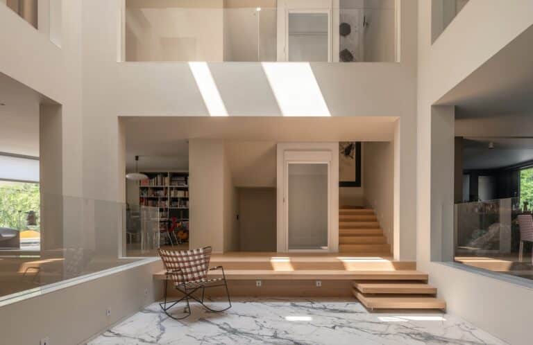 Interior view of the central light well in the House of Porous, showing a marble-floored courtyard, wooden steps, and glass railings with natural sunlight streaming from above in a multi-generational home.
