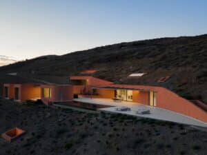 Exterior view of Oline resort in Syros at dusk, showing earth-toned buildings embedded in the rocky hillside with warm interior lighting.