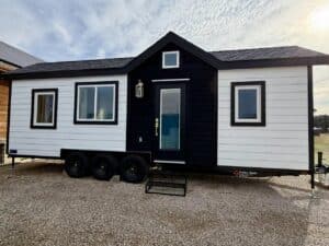 Exterior front view of the Betty tiny house on a triple-axle trailer, featuring white horizontal siding and black trim.