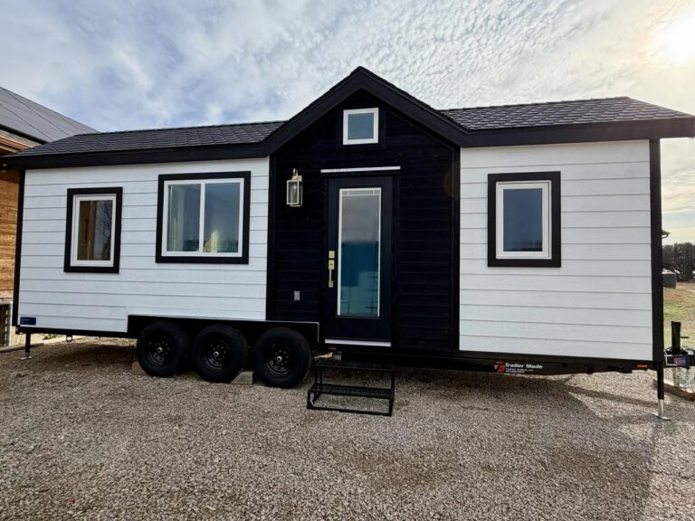 Exterior front view of the Betty tiny house on a triple-axle trailer, featuring white horizontal siding and black trim.
