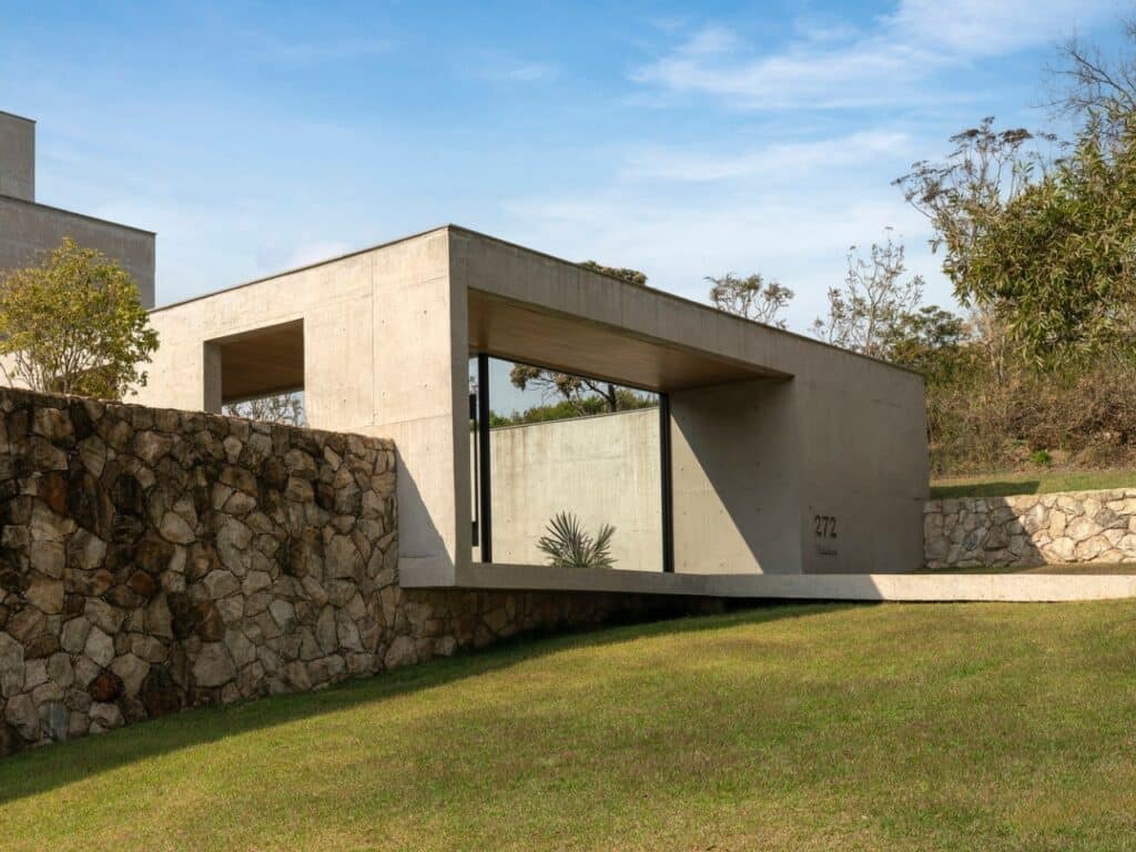 Modern concrete facade of GM House integrated with a natural stone wall on a grassy hill.
