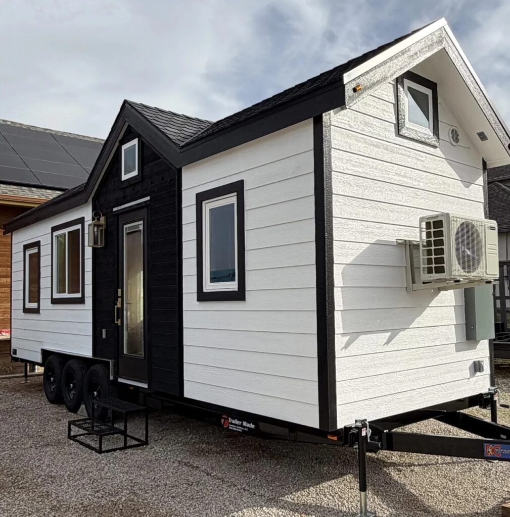 Side angle view of the Betty tiny house showing the entrance door, black-framed windows, and an external AC unit.