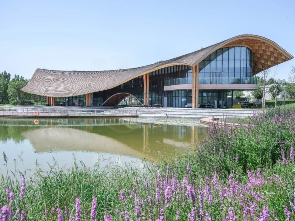 Exterior wide shot of the Silk Road Friendship Garden Restaurant featuring a flowing timber roof and glass facade reflecting on the lake.