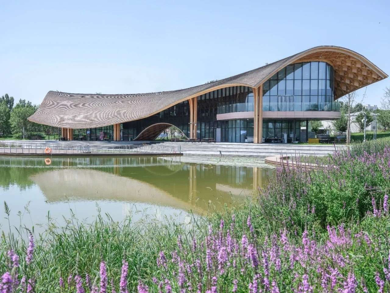 Exterior wide shot of the Silk Road Friendship Garden Restaurant featuring a flowing timber roof and glass facade reflecting on the lake.
