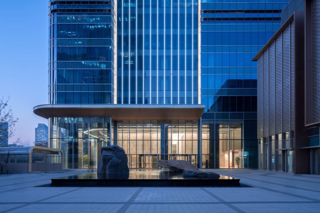 A dusk view of the Jinan Tower entrance in China, featuring illuminated vertical glass fins and a large natural stone sculpture on a dark water feature.