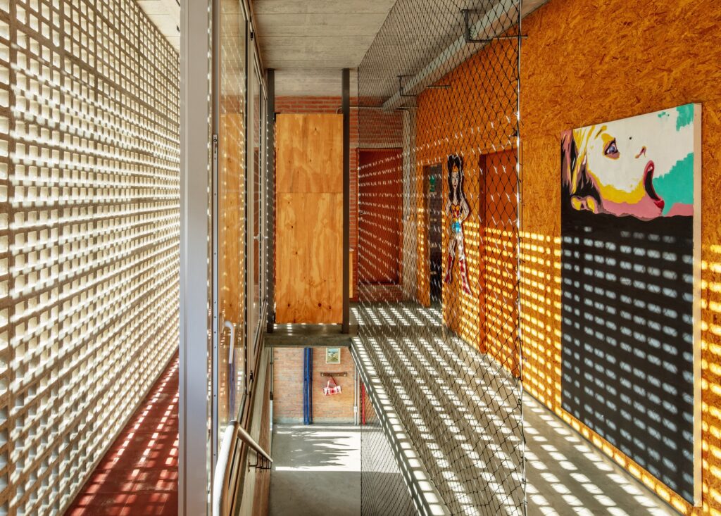 Upper floor corridor with sunlight filtering through a concrete cobogo wall and OSB wooden panels.