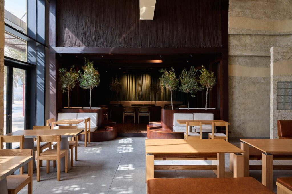 Spacious restaurant dining area with natural light, wooden furniture, and indoor trees against a dark wooden backdrop.