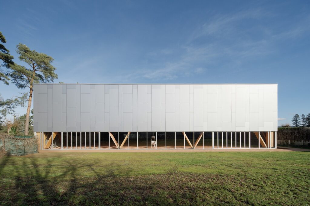 Symmetrical elevation of the Sporting Vichy sports hall under a clear blue sky, highlighting the timber structural supports behind glass.