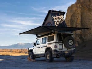 Side view of the Grand Bohemian 4x4 expedition vehicle with an expanded 270-degree awning and a rooftop pop-up tent in a desert landscape.