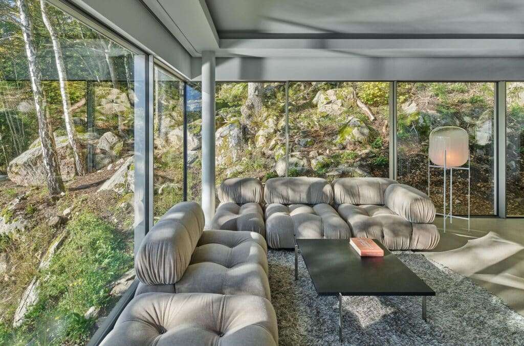 Interior living area of Bor&aring;s Getaway House featuring plush grey modular seating and large glass walls overlooking mossy rocks and forest trees.