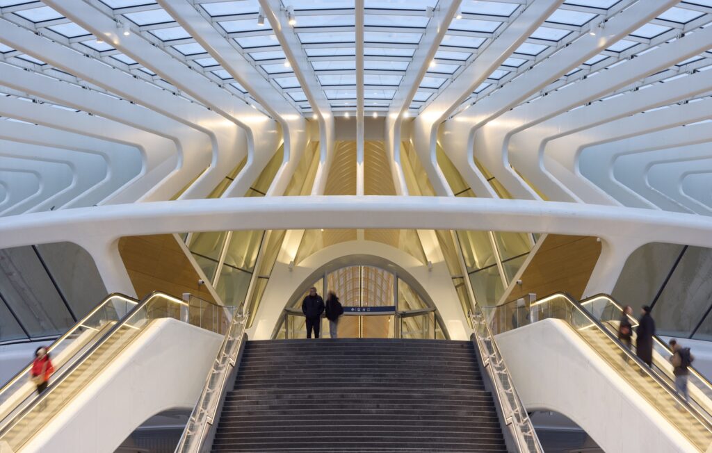 Modern wide staircase and escalators leading to the passenger gallery under the futuristic white steel canopy of Mons Station.