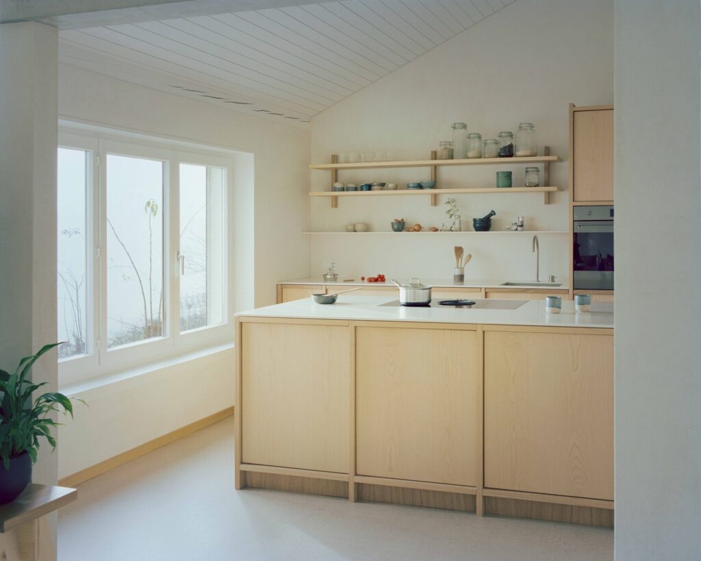 Close-up of a minimalist kitchen island with light ash wood cabinetry and open shelving against a white wall with a window.