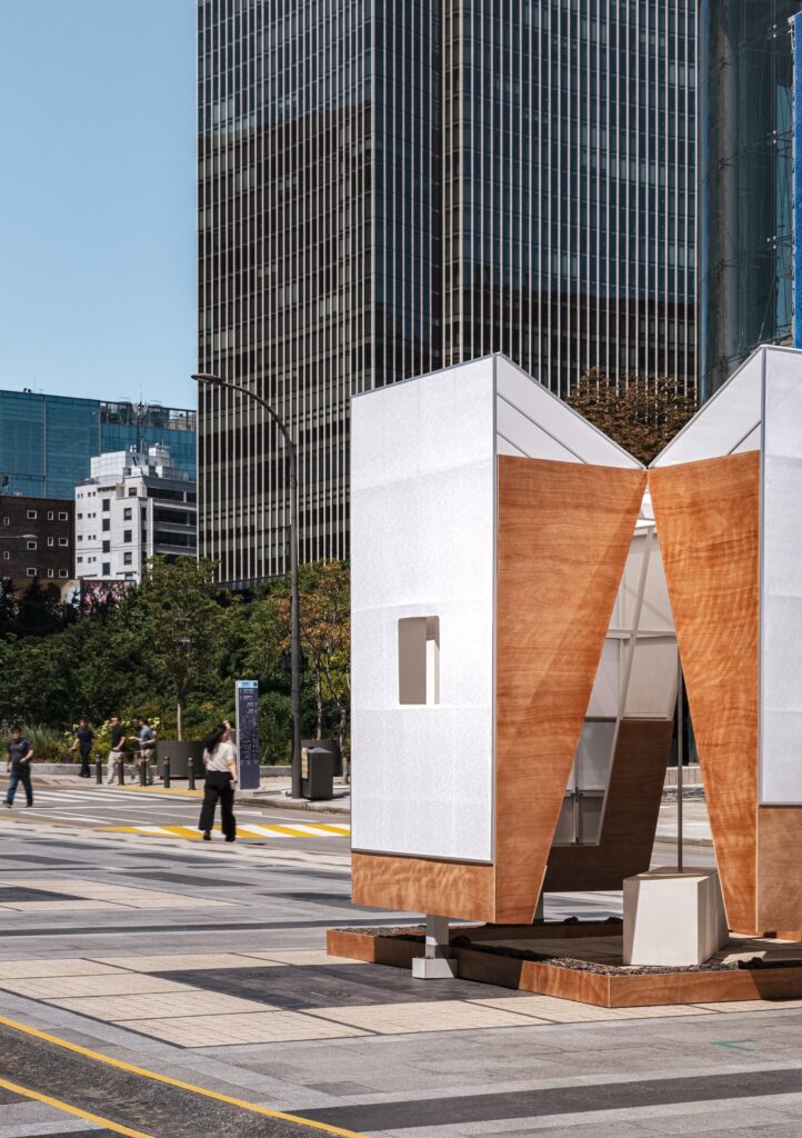 Side view of the TEUM pavilion exterior featuring white textured walls and triangular wooden panels on a paved public plaza.