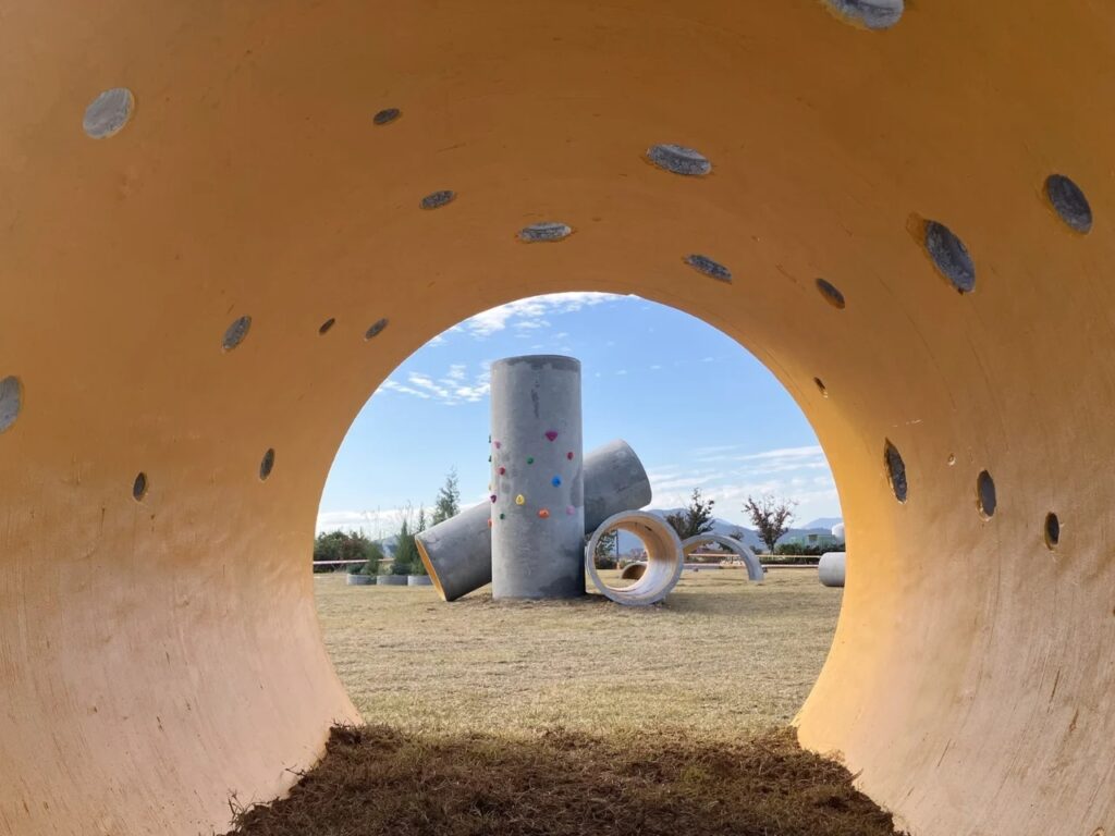A view from inside a concrete pipe looking out towards other cylindrical structures under a blue sky, showing yellow interior coating.