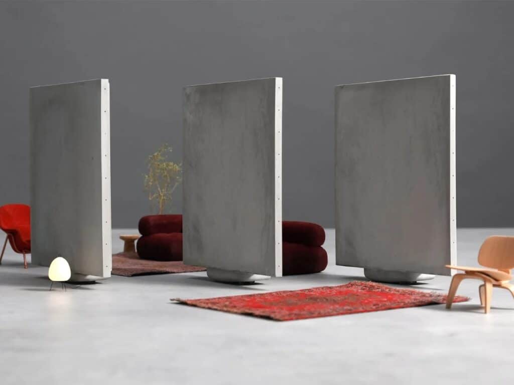 Three vertical concrete slabs standing independently in a minimalist living room setting with red chairs.