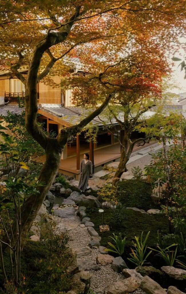 A woman walking through a lush Japanese garden with autumn maple leaves and a stone path leading to a traditional Kyoto house.