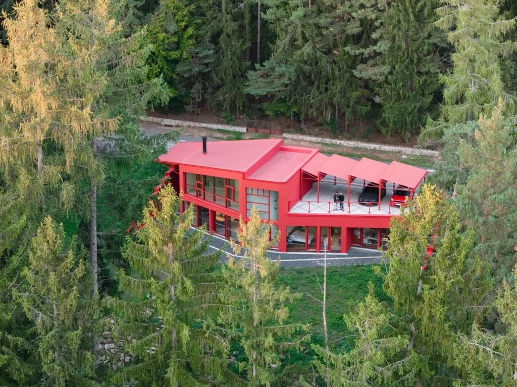 Aerial view of a bright red modern house with a steel structure nestled within a dense green forest in Jevany, Czech Republic.