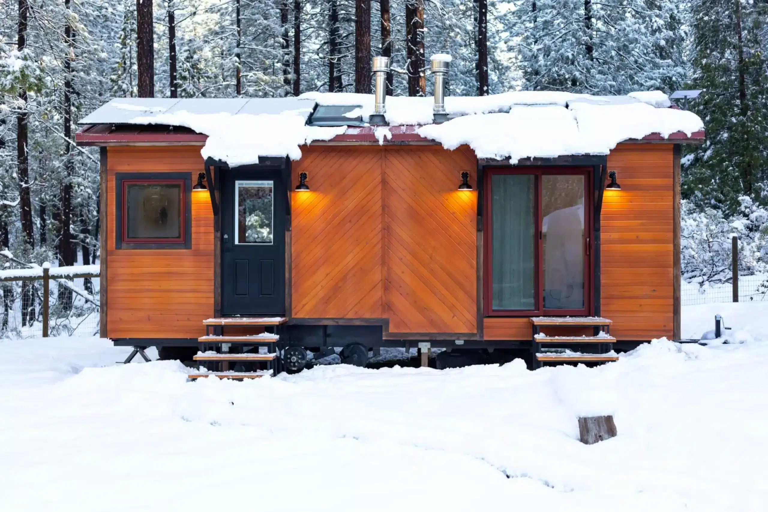 Exterior view of a wooden mobile double office on wheels settled in a snowy forest, featuring solar panels on the roof and warm outdoor lighting.