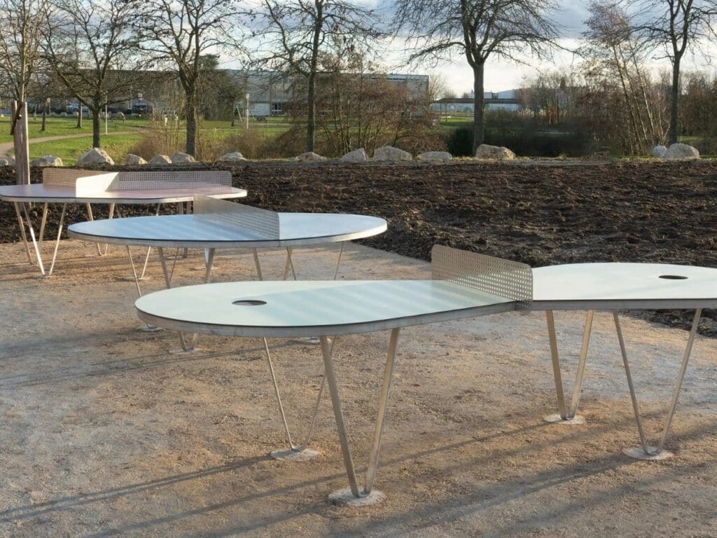 Three unconventional ping-pong tables installed in a public park setting, featuring circular and elongated shapes.
