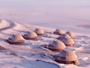 Aerial view of a cluster of volcanic-inspired architectural cabins in the snowy Inner Mongolia steppe at sunset.