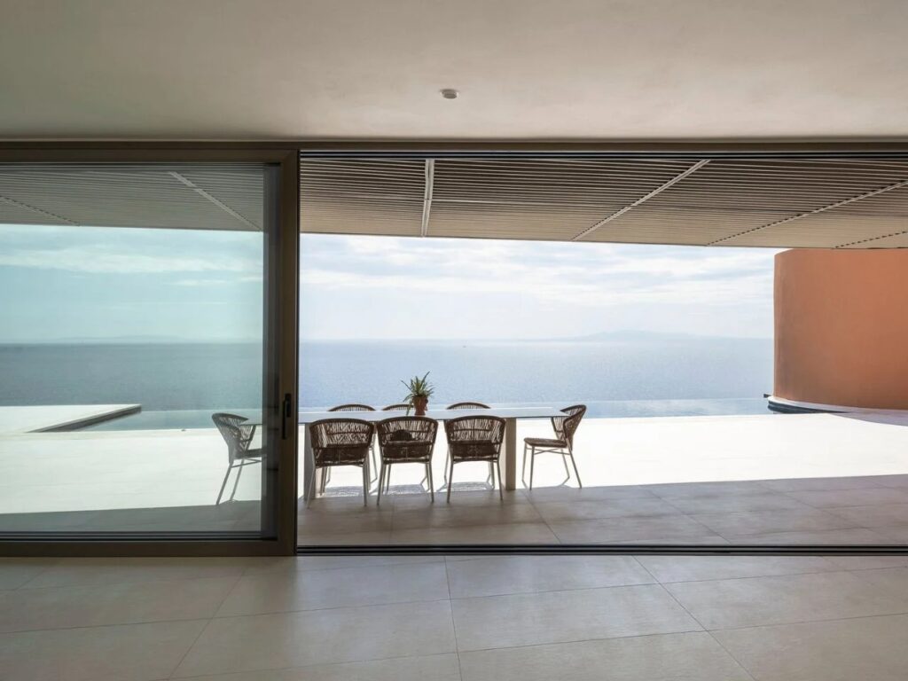 Interior view from the dining area looking out through large glass sliding doors to the Aegean Sea under a shaded pergola.