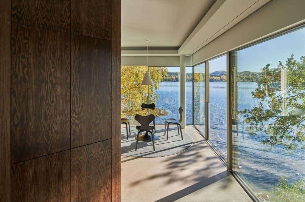 Dining area of Bor&aring;s Getaway House with a panoramic view of the lake through glass walls, featuring the dark wooden central core on the left.