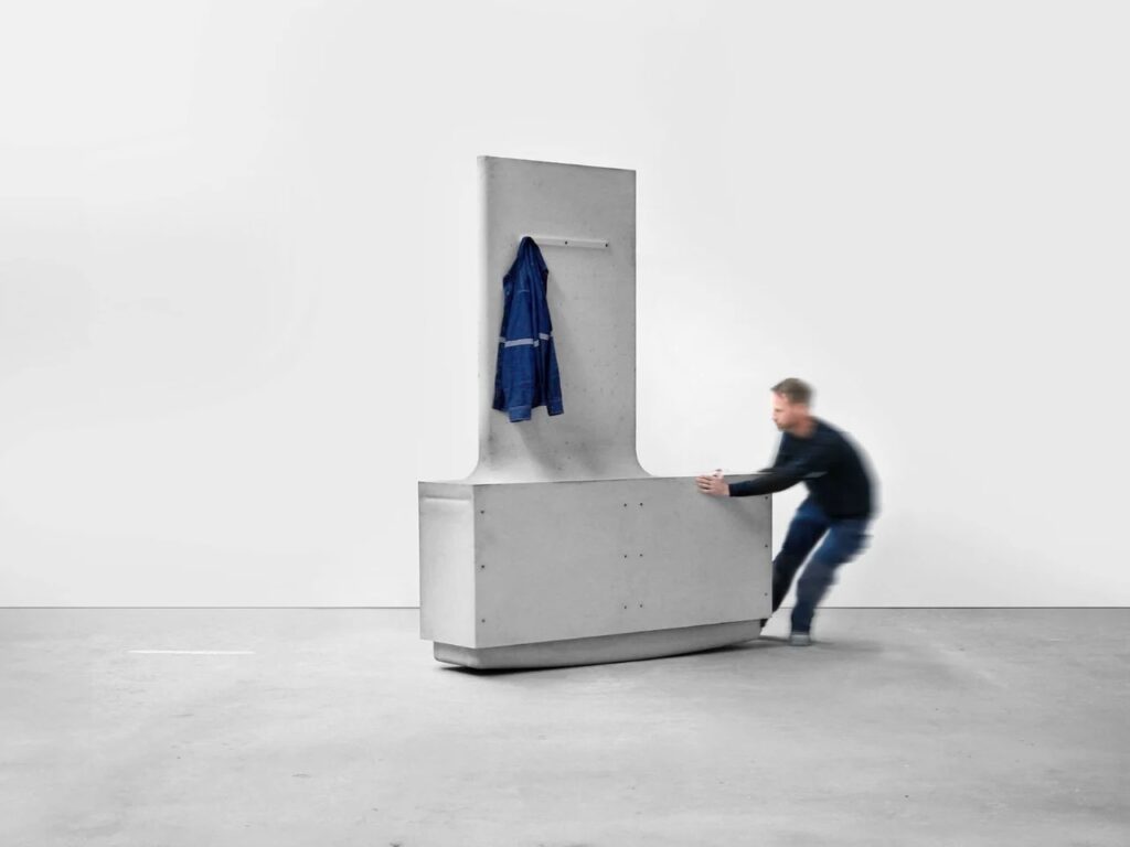 A blurred figure moving a tall concrete L-module with a jacket hanging on a peg, showcasing functional furniture integration.