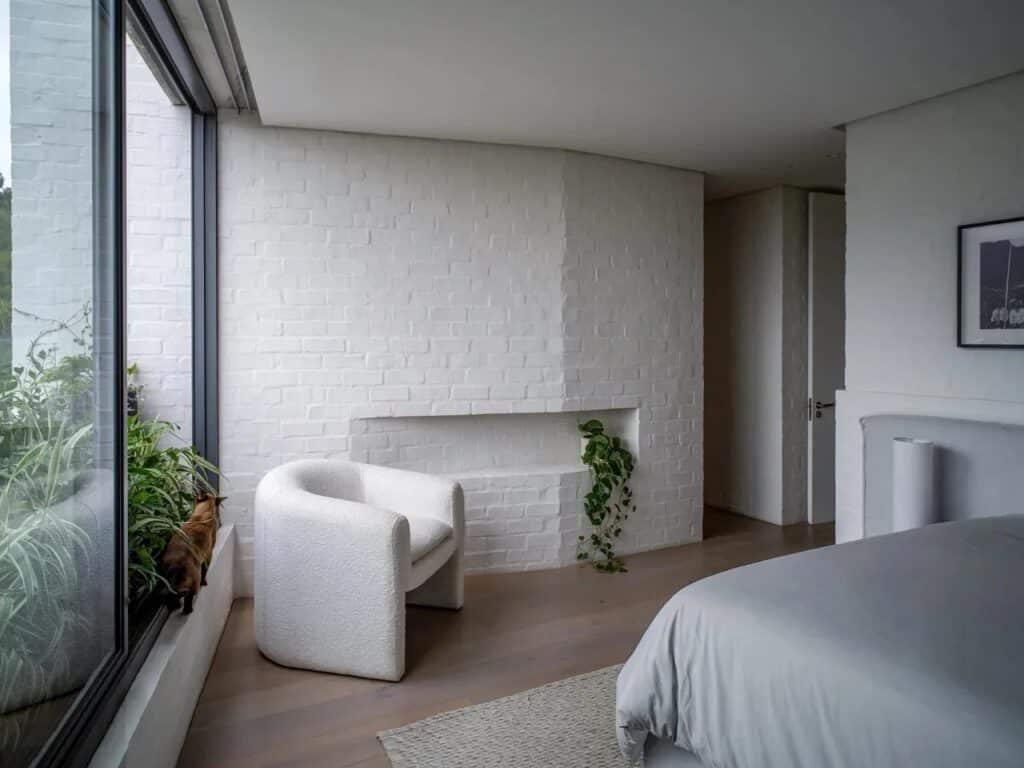 Minimalist white bedroom in Kenmore House with a large window overlooking lush greenery and a cozy white armchair.