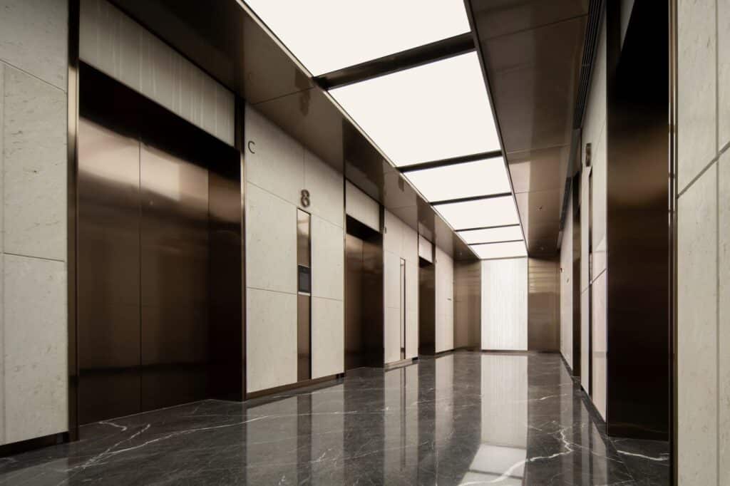 Interior view of an office floor lobby in Jinan Tower showing a minimalist hallway with bronze-toned elevator doors and light-colored marble walls.